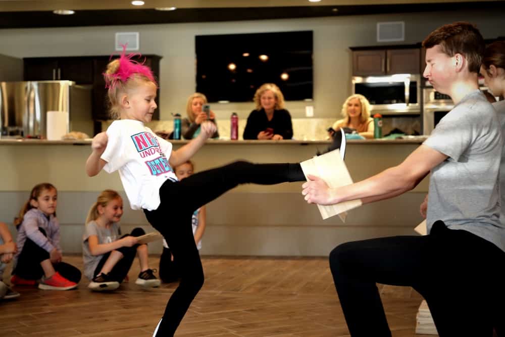 Self defense class, young woman in foreground kicking a board held by older boy